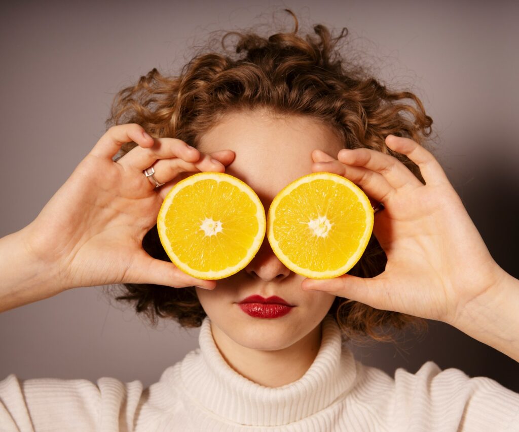 a blonde and curly haired american woman holding sliced orange fruit covering her 2 eyes