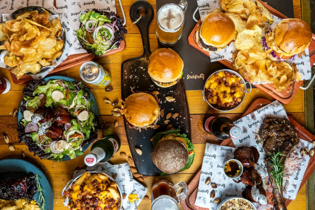 bunch of junk foods on table such as burgers. french fries, red meat steak, potato chips, alcohol, and soda laid to a wood table 