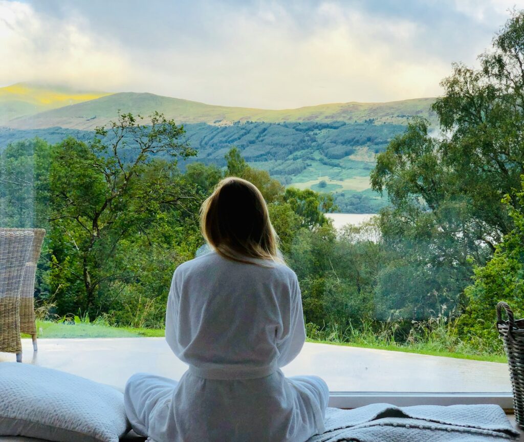 a short haired brunette woman wearing a white robe and sitting on bed looking at the mountains early morning