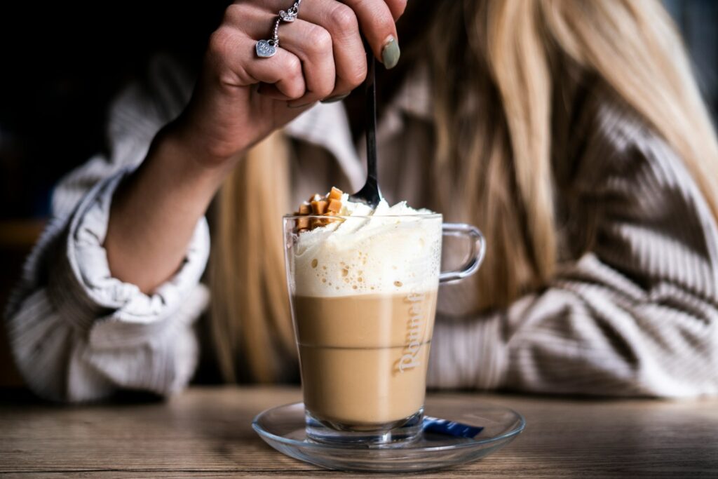 an american blonde woman drinking caramel macchiato coffee with whipped cream in a clear mug glass