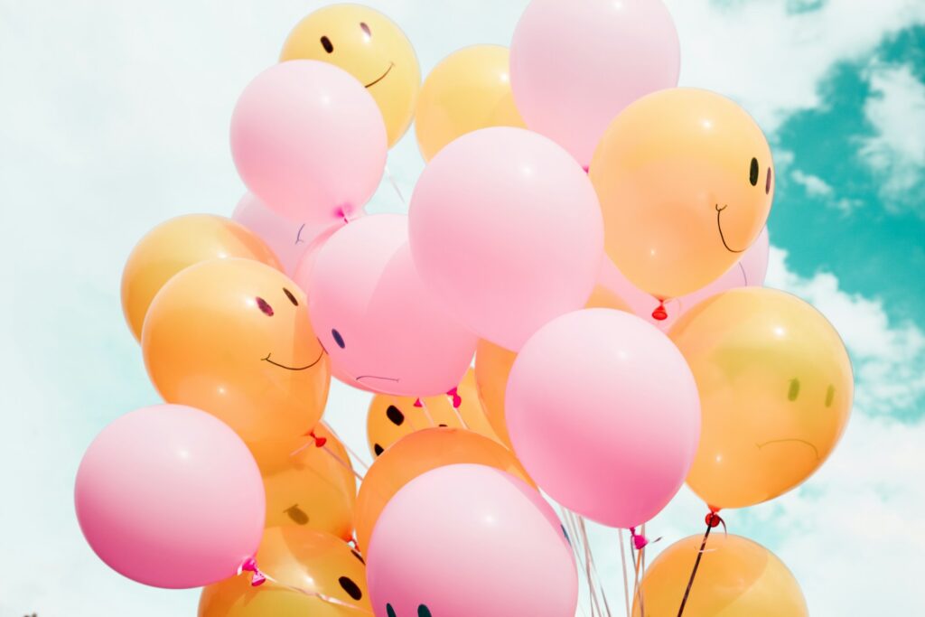 a bunch of mixed pink and orange balloons  with a drawn smiling faces floating up in the blue sky
