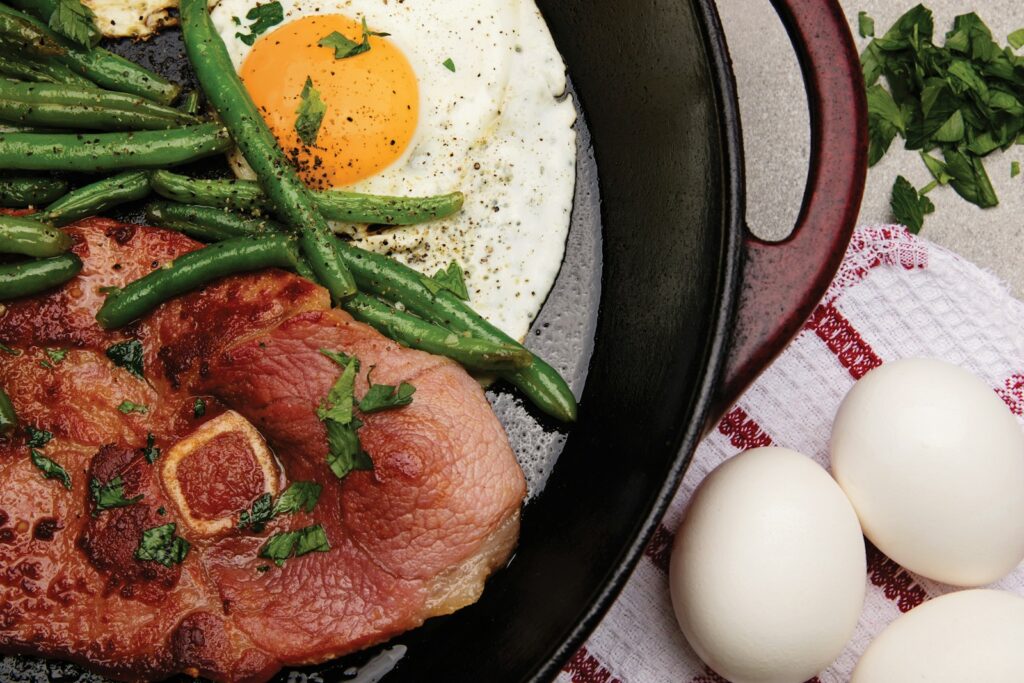 a fried egg with red meat and green string veggies cooked in a black skillet pan