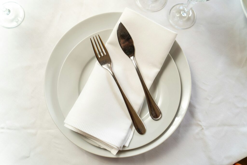 expensive plain dinner plate sets in ceramic pearl white with heavy metal spoon and fork placed on top of a table napkin